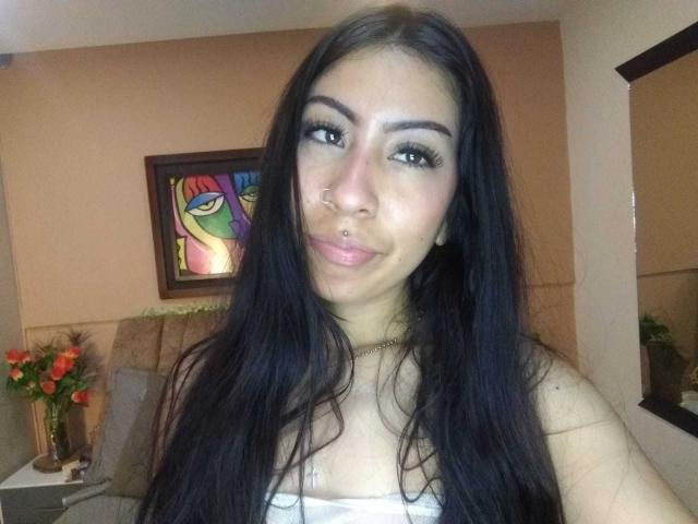 LinaSpearce with long black hair, brown eyes, nose ring, and lip piercing, wearing a white strap top, smiling slightly, in a room with colorful wall art and flowers.