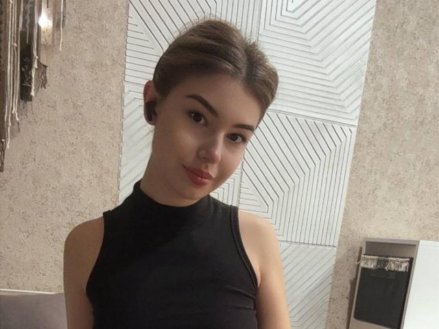 PearlineBerggren with short brown hair, wearing a black sleeveless top, small earbuds, slight smile, standing in a room, looking at the camera. She's got a cute face.