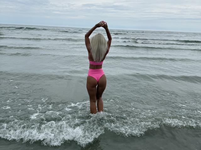 MoniqueBliss, con i capelli biondo platino, indossa un bikini rosa, è in piedi nell'acqua dell'oceano con le braccia alzate, mostrando i suoi glutei e la schiena toniche, i seni piccoli e la pelle bagnata..
