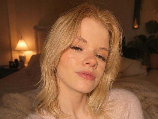 DonnaBellettiere ha i capelli biondi, la pelle chiara e le labbra rosa, indossa una maglietta bianca, con un leggero sorriso e una pelle morbida e luminosa, in una stanza accogliente e ben illuminata..