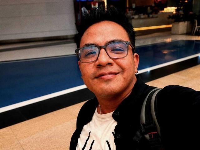 Rago420 a un visage rond, des cheveux noirs courts et porte des lunettes. Il porte une veste noire par-dessus une chemise blanche, il sourit et a un sac à dos. Ses fesses ne sont pas visibles..