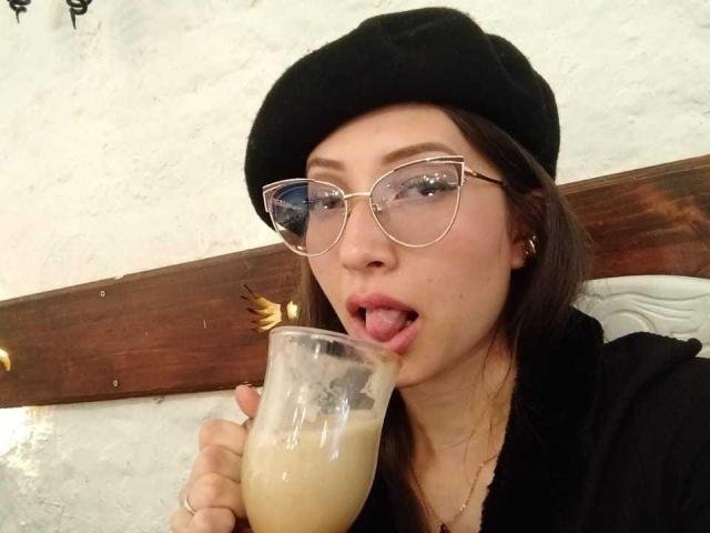 ElizabethMorrison with black beret, glasses, and gold hoop earrings, licking a frothy, light-colored drink from a glass, tongue out. Her black top contrasts with her light skin.