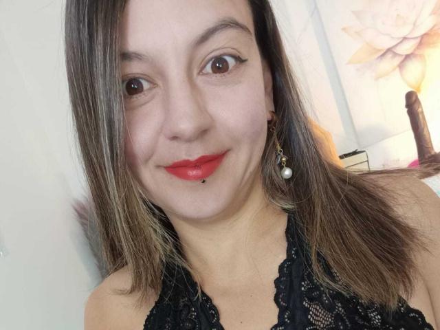 ArianaBlonde has light skin, brown eyes, and red lipstick. She's wearing a black lace halter top and a pearl earring, with medium brown hair and a cute smile.