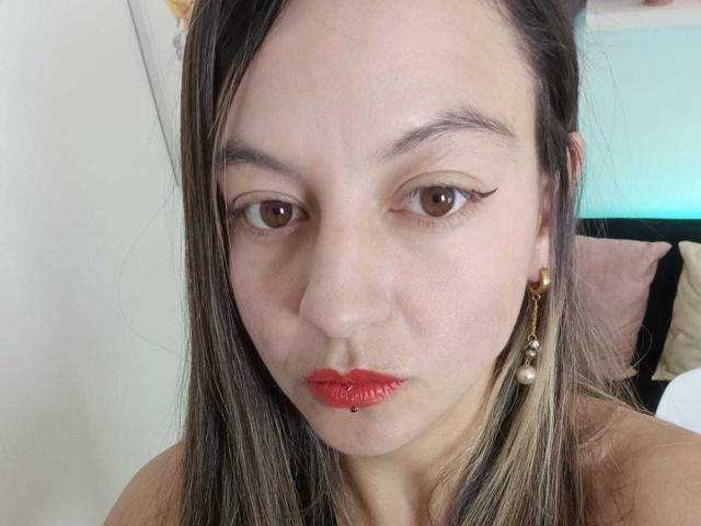 ArianaBlonde's face with red lipstick, straight brown hair, brown eyes, and gold earrings. She's pouting with a small silver lip piercing. Her tits are not visible.