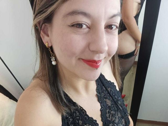 ArianaBlonde with red lipstick, black lace top, and earrings, showing a cute smile and pierced lower lip. Reflection shows her bare ass in black panties.