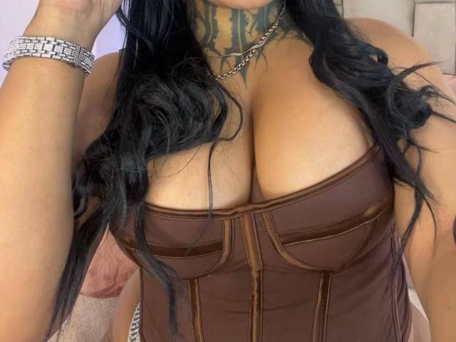 LairaHearvy's large, round tits in a brown, strapless corset, showing a tattooed neck and long black hair, with silver bracelets on her right wrist.