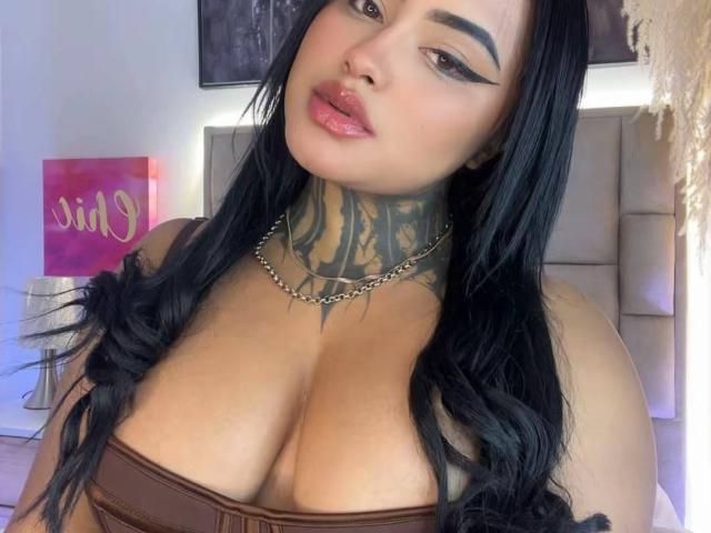 LairaHearvy has dark hair, thick eyebrows, and wears eyeliner with pink lipstick. She has large tits, a tattooed neck, and a chain necklace, looking down at the camera.