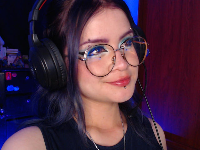 RavenThuwu with black headphones, round glasses, and a nose ring, has a lip piercing and a subtle smile, wearing a black sleeveless top. Blue and red lighting.