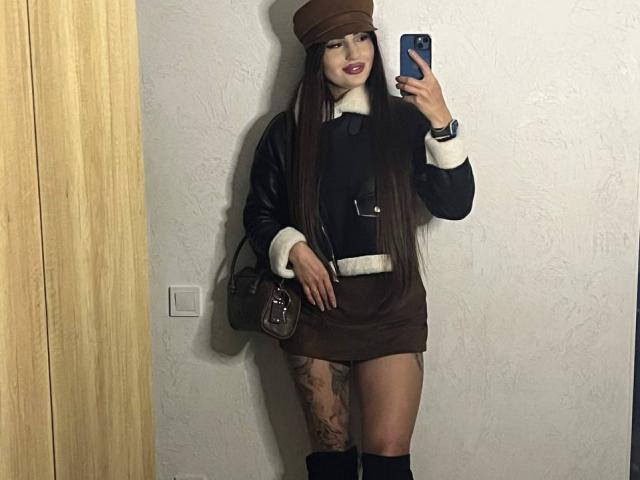Versavi with long brown hair, wearing a brown cap, black leather jacket, brown skirt, white fur trim, black boots, holding a phone, brown bag, tattoo on right thigh.