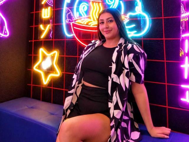 CatalinaRivero with dark hair, black crop top, black shorts, and a black-and-white patterned shirt, sitting on a blue bench, neon lights in the background, smiling.