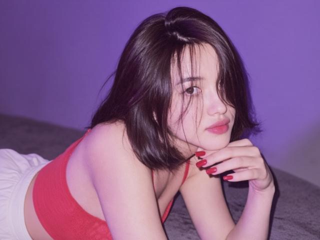MonikaMeel with short black hair, red bra, and red nails, lying on a gray surface, head resting on her hand, looking at the camera with a slight smile.