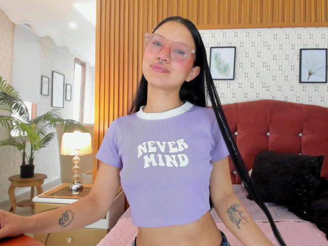 SophieDelaney with long black braids, pink glasses, and a purple crop top, showing her small tits and tattoos, smiling in a bedroom with a red headboard and lamp.