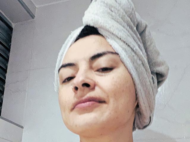 KatParadise with a gray towel wrapped around her head, smiling slightly, showing a few pimples on her face, in a bathroom setting. Her nose is small and she's cute.
