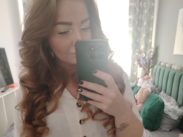 EveBliss with long, wavy brown hair, white button-up shirt, holding green phone, taking mirror selfie, showing black nail polish, small tattoo on right hand, subtle smile, soft lighting.