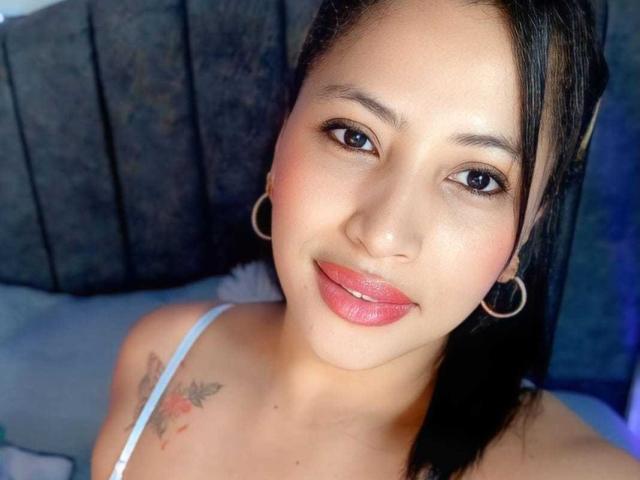 RoxaneMilf has medium skin, black hair, and hoop earrings. She's smiling with pink lipstick, showing a small tattoo on her right shoulder, and wearing a white top.