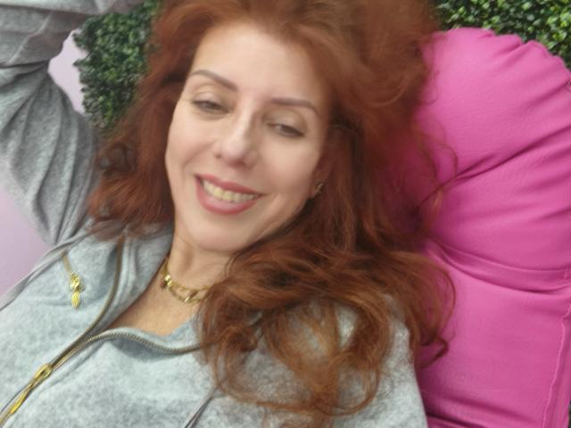Khharha with red hair, smiling, wearing a gray zip-up hoodie, and a gold necklace, lying on a pink pillow, has a cute face and medium-sized tits.