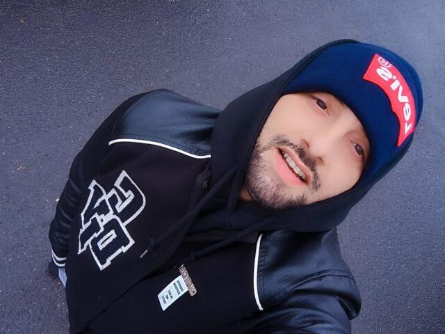 TommyDum in a black hoodie with white lines, blue cap with red "SEEN" tag, leather jacket, smiling with a beard, hands in pockets, standing on wet pavement.