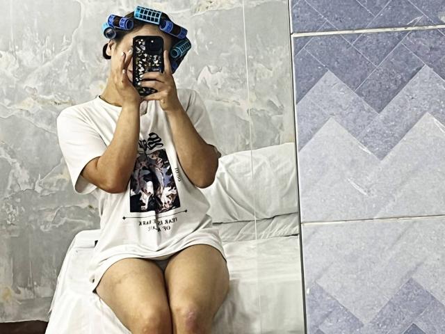ValentineDufour sitting on a bed, wearing a white tee with a black graphic, hair in blue curlers, holding a phone with a patterned case, covering face.