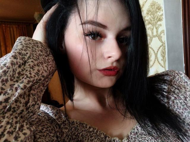 MissCourtney with black hair, red lipstick, and a floral patterned top, looking at the camera with one hand in her hair. She has a pretty face and long eyelashes.