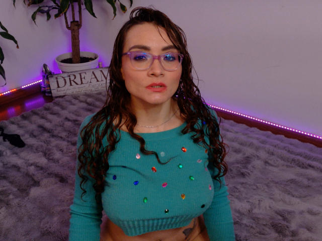 Drikka with wet brown hair, purple glasses, and a teal crop top with colorful buttons, sitting on a grey fur rug, showing her boobs. Purple LED strip lights in background.