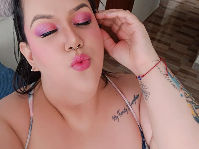 GiiannaMorrys with pink eyeshadow, black eyeliner, nose ring, pink lips, black hair, tattoo on right shoulder, tattoo on right wrist, hand near face, wearing a pink strap top.