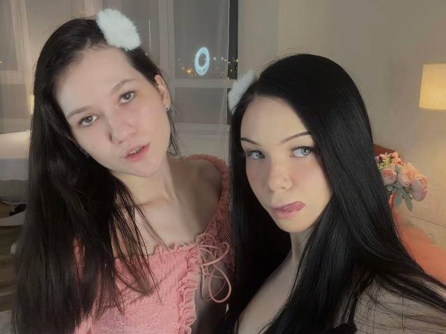 LornaAndGerry with long black hair, wearing pink lace-up top and white furry cat ears, and Gerry with black hair, pink lipstick, and black top, both with cute expressions.