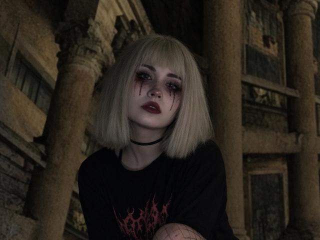 EmelyDilsaver has blonde bob, bloody eyes, dark lipstick, black choker, black shirt with pink design, fishnet arm, standing against ancient columns, looking sad and tired.