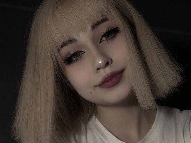 EmelyDilsaver with blonde bob, dark eyeliner, long lashes, pink lips, small beauty mark on left cheek, white shirt, looking at camera, soft lighting, slightly smiling, cute face.