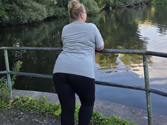 RadaRori's ass is up in black pants, standing with back to camera, leaning on a green metal railing by a reflective waterway, wearing a grey shirt, blonde bun.