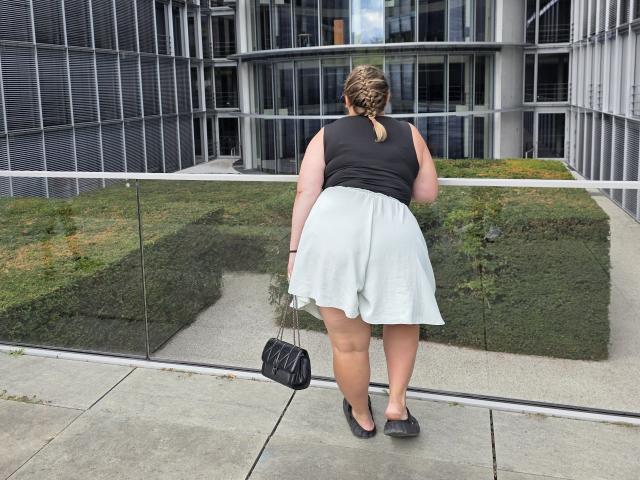RadaRori, with a chubby ass in a black sleeveless top and white skirt, stands with back to camera, holding black purse, in front of a glass wall, outside.