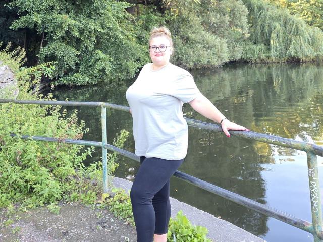 RadaRori stands by a metal railing, wearing a gray shirt, black leggings, glasses, and a watch, with her hand on the railing, by a reflective waterway.