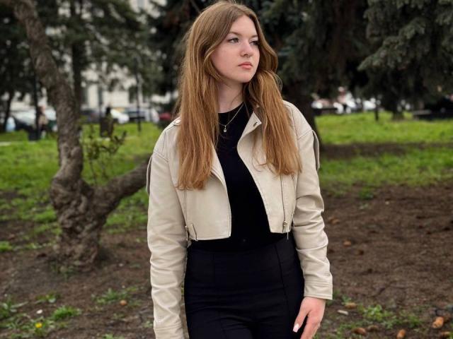 Conchita Mosburg, con lunghi capelli castani, indossa una giacca di pelle beige, una maglietta nera e pantaloni neri, è in piedi in un parco e guarda di lato con un'espressione neutra..