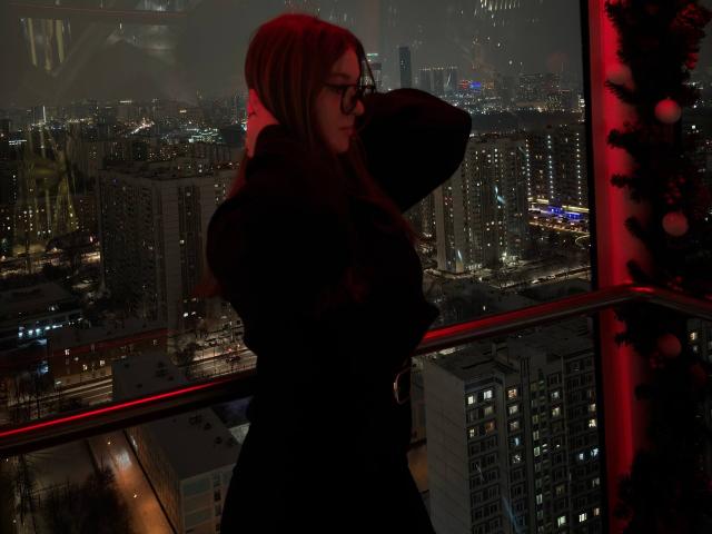 Conchita Mosburg mit roten Haaren, Brille, schwarzem Mantel und schwarzem Hut, stehend vor einem dunklen Stadtfenster, rotes Licht auf der rechten Seite, städtische Nachtansicht, Blick zur Seite..