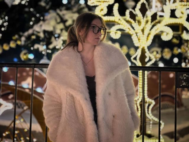 ConchitaMosburg with glasses, wearing a fluffy white fur coat, standing in front of a black metal railing, looking to the side, brown hair in a ponytail, night setting.