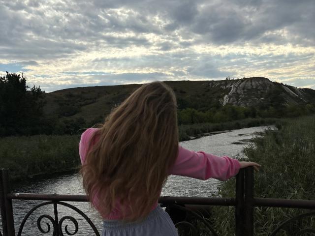 ConchitaMosburg with long, wavy brown hair, pink long-sleeve shirt, and white skirt, leans on a metal railing by a river, back to the camera, under cloudy sky.