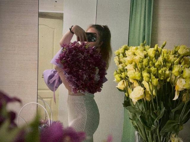 Conchita Mosburg, avec un bouquet violet recouvrant ses seins, portant une robe blanche, moulante et en dentelle, prenant un selfie devant un miroir, entourée de roses jaunes et de fleurs violettes..