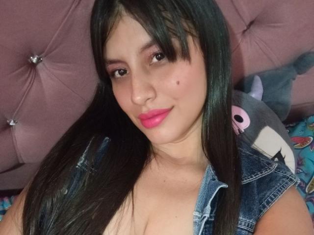 MadissonFontaine with long black hair, pink lipstick, and denim shirt, showing cleavage, smiling at the camera, pinkish-purple tufted headboard in the background.