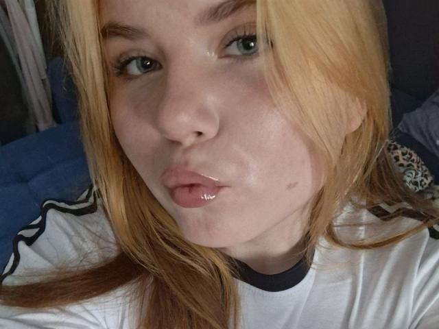 LatashaRiney with glossy, shiny lips, pouty, and a small beauty mark on her left cheek. She has blonde hair, green eyes, and a white shirt with black trim.