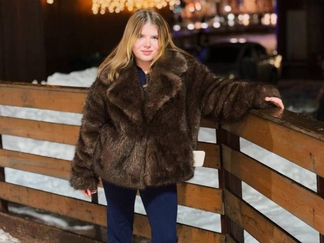 LatashaRiney with long blonde hair, wearing a dark brown fur coat and blue pants, leaning on a wooden fence, smiling. Her tits are not visible.