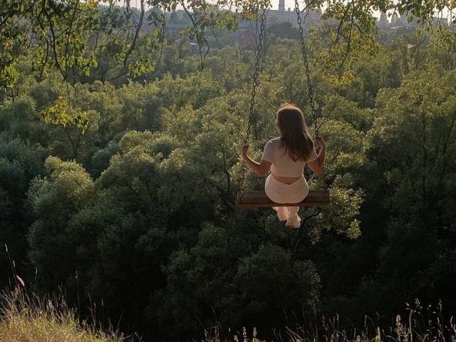 Latasha Riney, con el trasero expuesto, está sentada en un columpio de madera, agarrando las cadenas, mirando hacia un bosque verde, la luz del sol se filtra entre las hojas, de espaldas a la cámara, cabello castaño, vestida con ropa interior blanca..