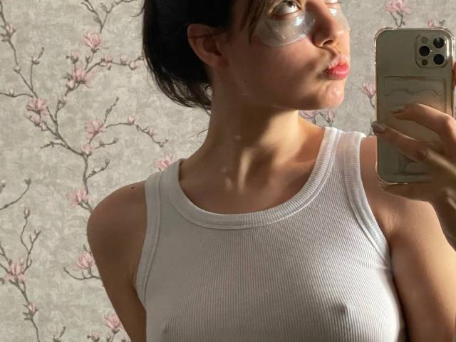 HortenseSpruit with a white ribbed tank top, glossy face mask, pouty lips, and dark hair in a ponytail, holding a gold phone, showing her tits and ass.