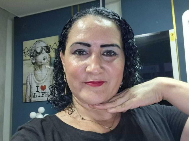 SilverCool69 ha i capelli neri ricci, il rossetto rosso e indossa una maglietta nera con una collana d'oro. Ha il mento appoggiato sulla mano e sta guardando la fotocamera..