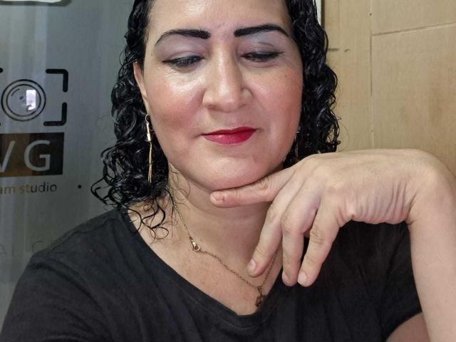 SilverCool69 heeft krullend zwart haar, rode lippenstift en een lichte glimlach. Ze draagt een zwarte top, een gouden ketting en oorringen, en haar hand rust op haar wang..