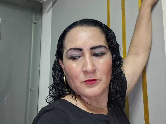 SilverCool69 con cabello negro rizado y húmedo, sombra de ojos morada, lápiz labial rojo, camisa negra, brazo levantado, aretes dorados y una ligera sonrisa, mostrando un poco de su rostro sudado..