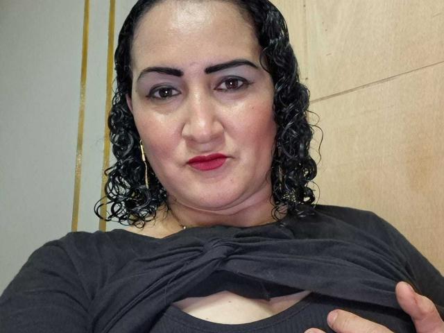 A SilverCool69 tem cabelo preto encaracolado, sombra roxa, batom vermelho e está levantando a camisa preta para expor seus pequenos seios pálidos. Ela está usando brincos de argola..