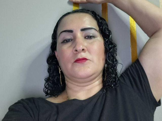 SilverCool69, una donna corpulenta con capelli neri ricci, rossetto rosso, una maglietta nera e orecchini a forma di croce dorata, è appoggiata a un palo giallo, alzando il braccio destro..
