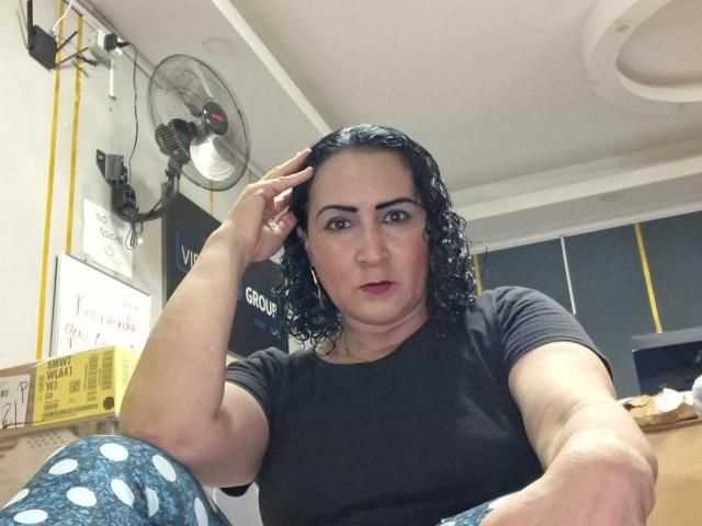 SilverCool69 con capelli neri ricci, rossetto rosso, camicia nera, pantaloni blu a pois, una mano tra i capelli, espressione seria, seduto in un ufficio, ventilatore a parete, luce a soffitto, scrivania di legno..