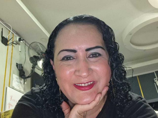 A SilverCool69 tem cabelo preto encaracolado, sobrancelhas escuras, batom vermelho e está sorrindo com a mão no rosto. Ela está vestindo uma camisa preta, parecendo feliz e atraente..