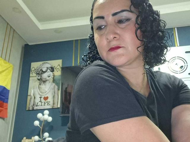 SilverCool69, avec des cheveux noirs bouclés, du rouge à lèvres et un t-shirt noir, a l'air légèrement agacé. L'arrière-plan comprend une peinture d'une femme avec le texte "I ♥ LIFE"..