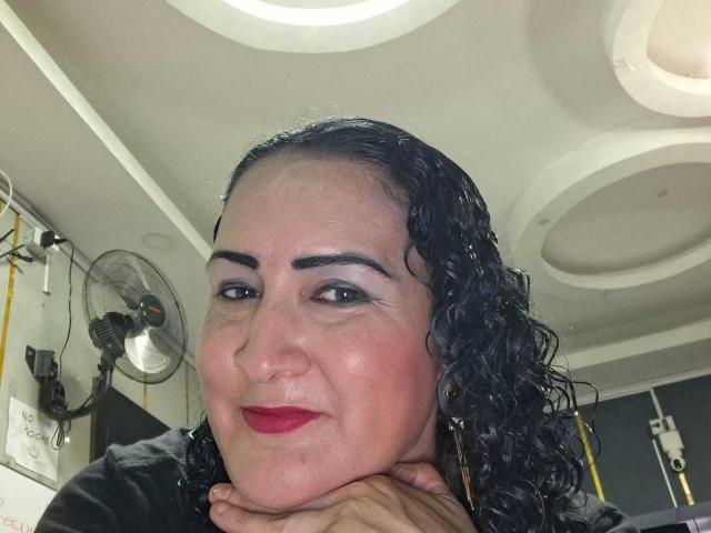 SilverCool69 tiene el pelo negro y rizado, lápiz labial rojo y maquillaje de ojos oscuro. Está sonriendo, con el mentón apoyado en la mano, y lleva una blusa negra. Su piel es de tono medio..